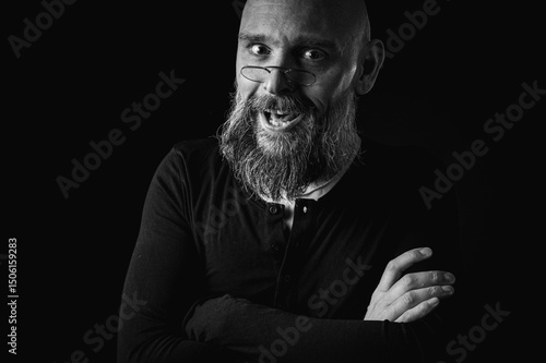Black and white close-up portrait of a funny emotional weird bald man with a long gray beard and glasses making a funny face. Concept of eccentricity, humor and expressive personality.