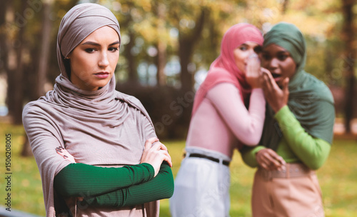 Bullying. Fake Friends Talking About Unhappy Muslim Woman Standing Alone In Park Outdoor. Female Friendship Problem, Outcast Person Concept. Selective Focus