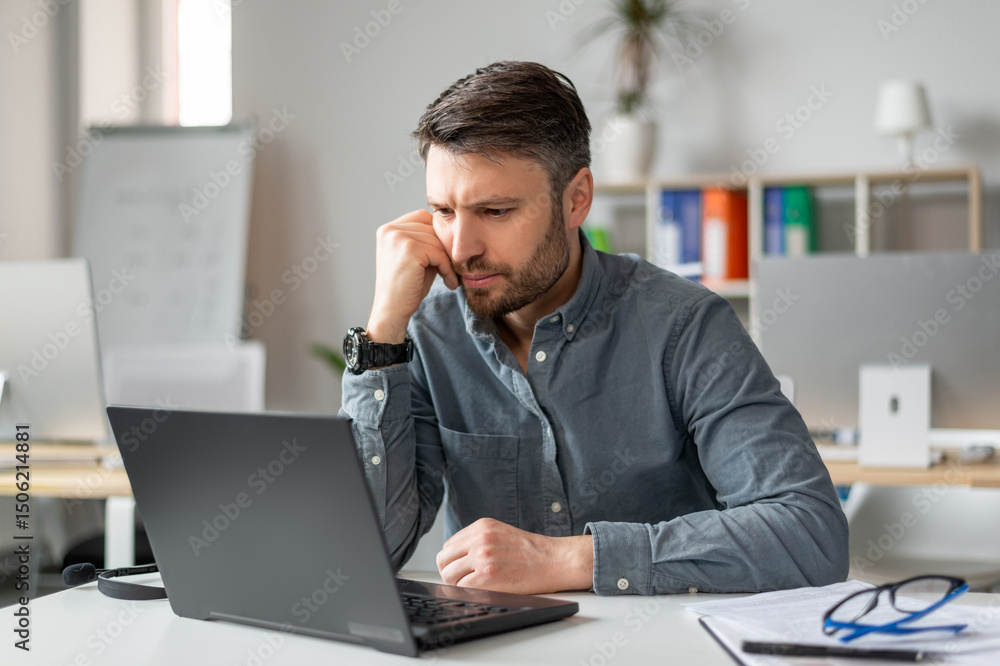 Serious middle aged businessman using laptop computer and reading reports, sitting at workplace in office. Business and entrepreneurship problems concept Serious middle aged businessman using laptop computer and reading reports, sitting at workplace in office. Business and entrepreneurship problems concept