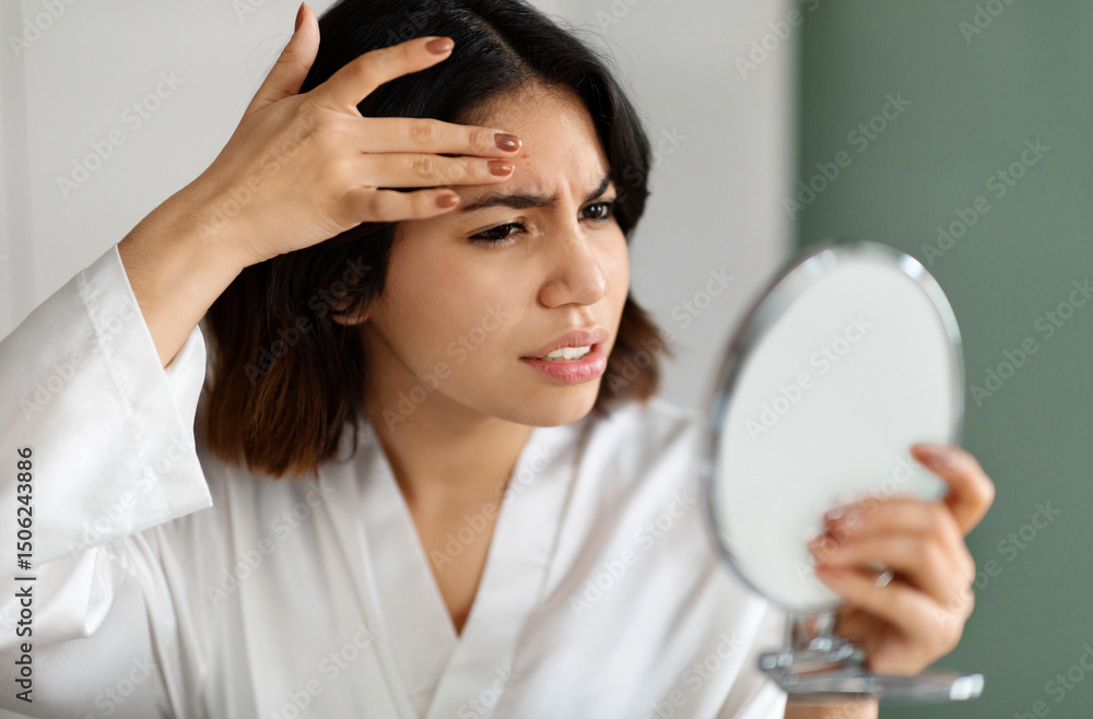 Naklejka premium Upset arabic young woman in white silky robe looking at hand mirror, checking her skin, touching her forehead with red pimple, home interior, closeup photo. Acne, pimples, dull skin