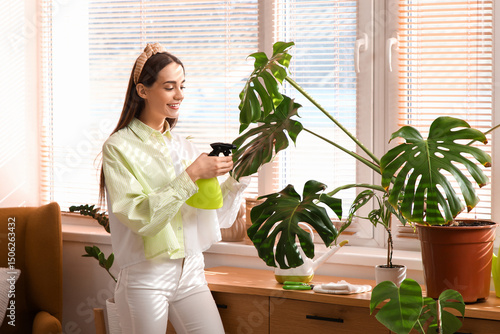 Beautiful young woman spraying water on houseplant at home