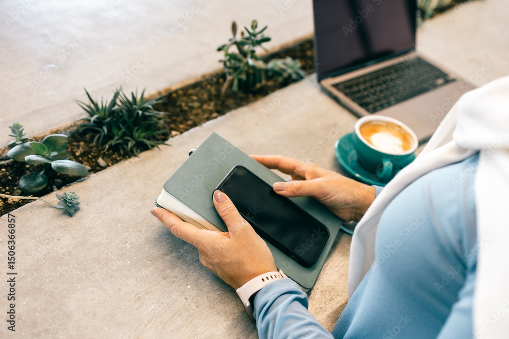 Obraz premium Close-up of woman holding smartphone and notebook in cafe with coffee and laptop