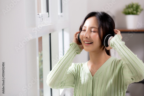 Portrait happy young woman wearing headphones listening music while standing with fun and enjoy in the living room at home, happiness female with entertainment, one person, lifestyle concept.
