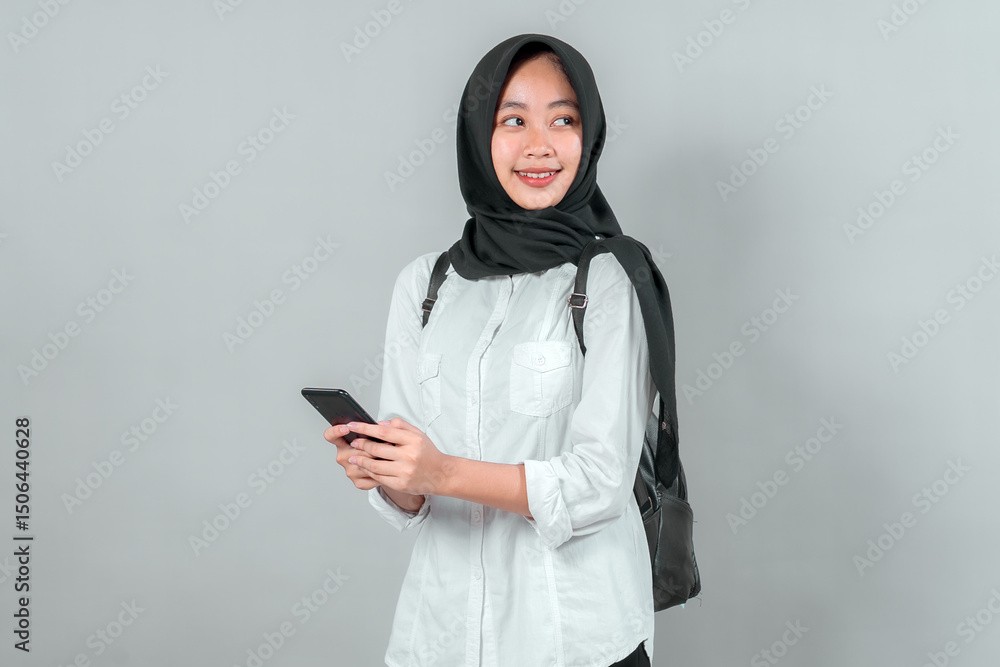 Cheerful Muslim schoolgirl wearing a hijab and backpack holding a smartphone while smiling and looking away. Confident student ready to start the new academic year with enthusiasm. Cheerful Muslim schoolgirl wearing a hijab and backpack holding a smartphone while smiling and looking away. Confident student ready to start the new academic year with enthusiasm.