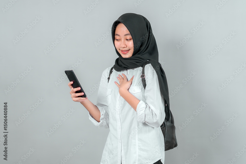 Smiling Muslim schoolgirl in black hijab and white shirt, wearing a backpack and holding a smartphone, looking at the screen while gently touching her chest, ready for school. Smiling Muslim schoolgirl in black hijab and white shirt, wearing a backpack and holding a smartphone, looking at the screen while gently touching her chest, ready for school.