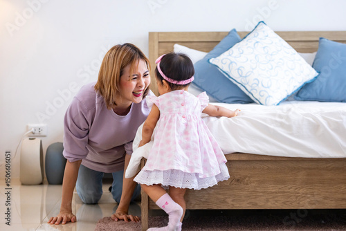 Mother is playing peek a boo or hide and seek with her baby toddler in the bedroom while the girl is laughing in happiness for good parenting and healthy raising kid