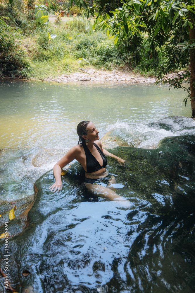 Fototapeta premium woman enjoys a serene moment while sitting in a clear, natural pool, surrounded by vibrant green foliage. peaceful water flows gently over smooth rocks under the warm sun