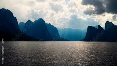 Dramatic mountain range silhouette under stormy sky with sun rays shining through dark clouds over still lake waters