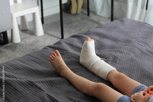 Child resting on a bed with a cast on one leg indicating a recent injury while other foot remains uninjured. Boy with broken leg at home.
