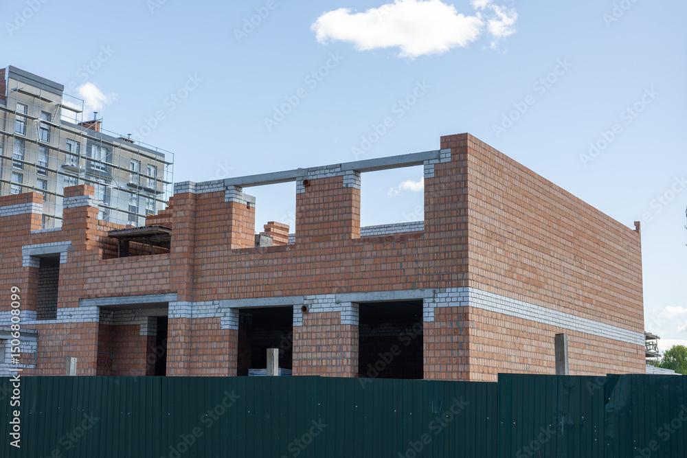 Obraz premium Brick residential building under construction behind green fence on clear sky background. 