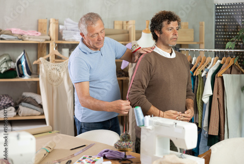 Senior man tailor takes measurements from senior male client, using tape measure, measures body size for custom-made dress..