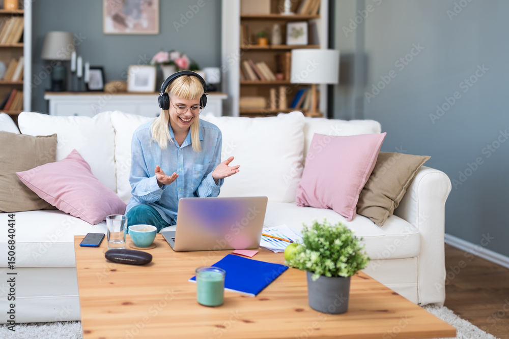 Young woman wearing headphones having video call on laptop, sitting on cozy couch at home, working remotely or studying online, gesturing while talking, surrounded by coffee, notes, and documents. Young woman wearing headphones having video call on laptop, sitting on cozy couch at home, working remotely or studying online, gesturing while talking, surrounded by coffee, notes, and documents.