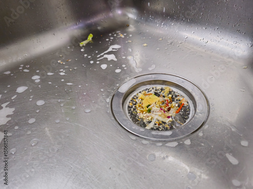 Fototapeta Close-up of a stainless steel kitchen sink with food scraps collected in a mesh drain strainer, surrounded by soap suds and water droplets