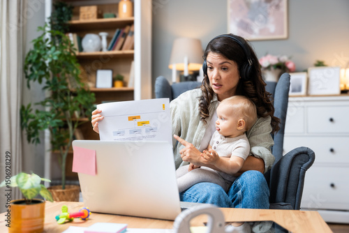 Single mother with baby on lap speaks emotionally via laptop video call to ex-husband about overdue child support, alimony, sitting in armchair wearing wireless headphones, facing financial stress. photo