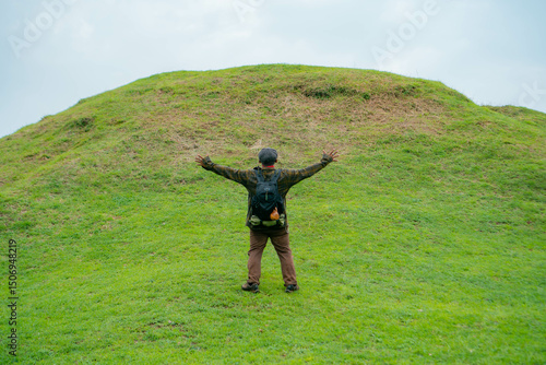 A male adventurer is enjoying the beauty of the expanse of green grass. The Asian male traveler relaxes and is amazed by the natural charm of the clean and beautiful green hills.