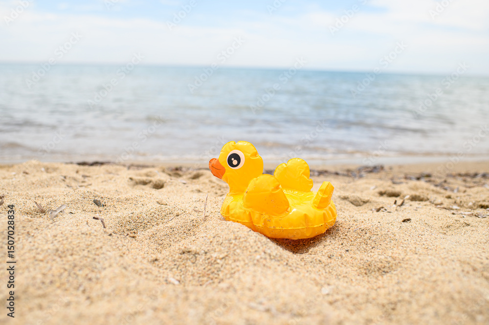 Obraz premium Yellow inflatable duck toy lying on sandy beach near the sea. Bright summer concept, vacation vibes, seaside fun, minimal beach background with copy space.