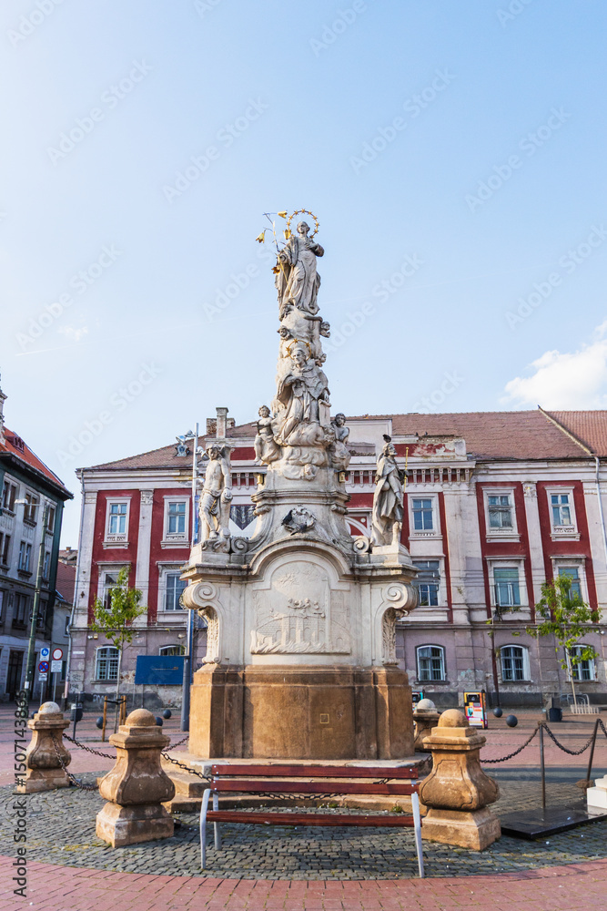 Obraz premium Detailed view of a tall, ornate stone monument adorned with multiple figures and sculptures, standing prominently in Timisoara's historic Liberty Square, Romania.