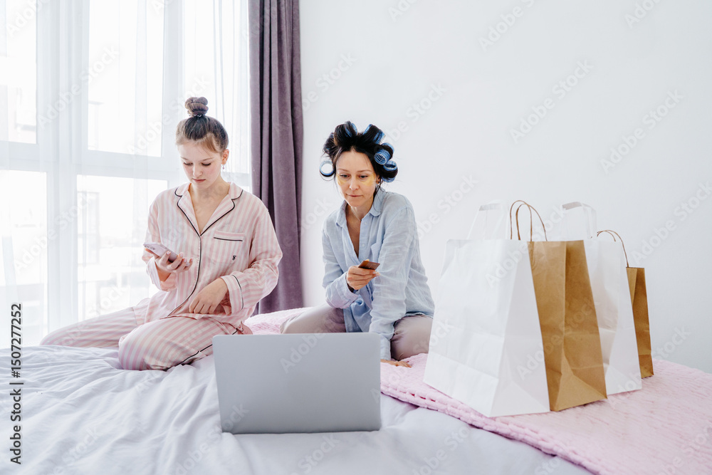 Obraz premium Teenage girl and mother in her 40s lying on bed in bright bedroom, browsing laptop during online shopping. Paper bags nearby. Morning routine showing digital habits and connection