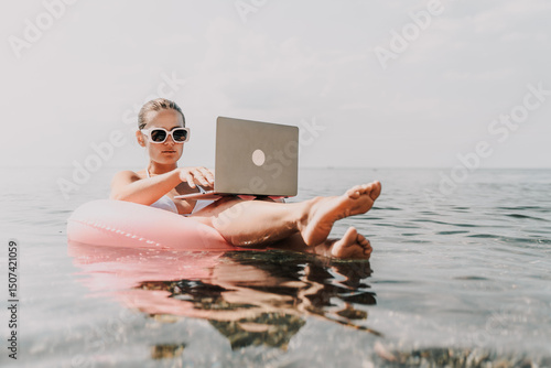 Canvas Print A woman is sitting on a pink inflatable raft in the ocean, using a laptop
