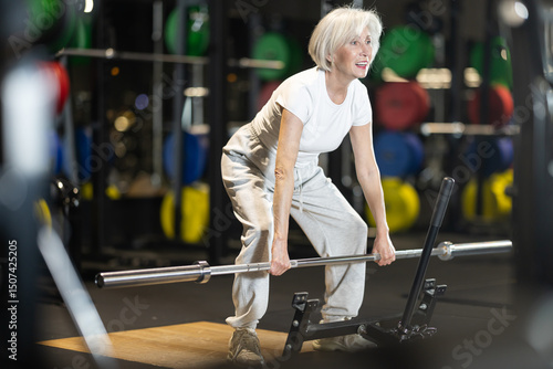 Fotografia Senior woman trains muscles of arms, standing up doing barbell exercises, differentiated weighting in crossfit gym
