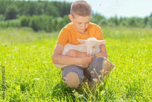 A young boy in an orange shirt sitting in a field embracing a white goat