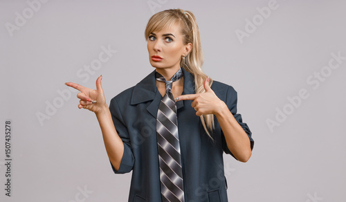 A woman with blonde hair in a half-updo, wearing a navy blazer and a striped tie, points with her left hand while looking to the side.