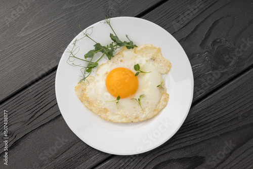 A sunny side up egg with fresh micro greens on a white plate over dark wood.