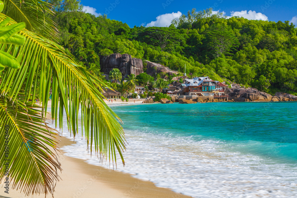 Fototapeta premium Sandy beach with palms and blue water of the Indian Ocean and volcanic stones with a hotel in the background. Island Mahe, the Seychelles