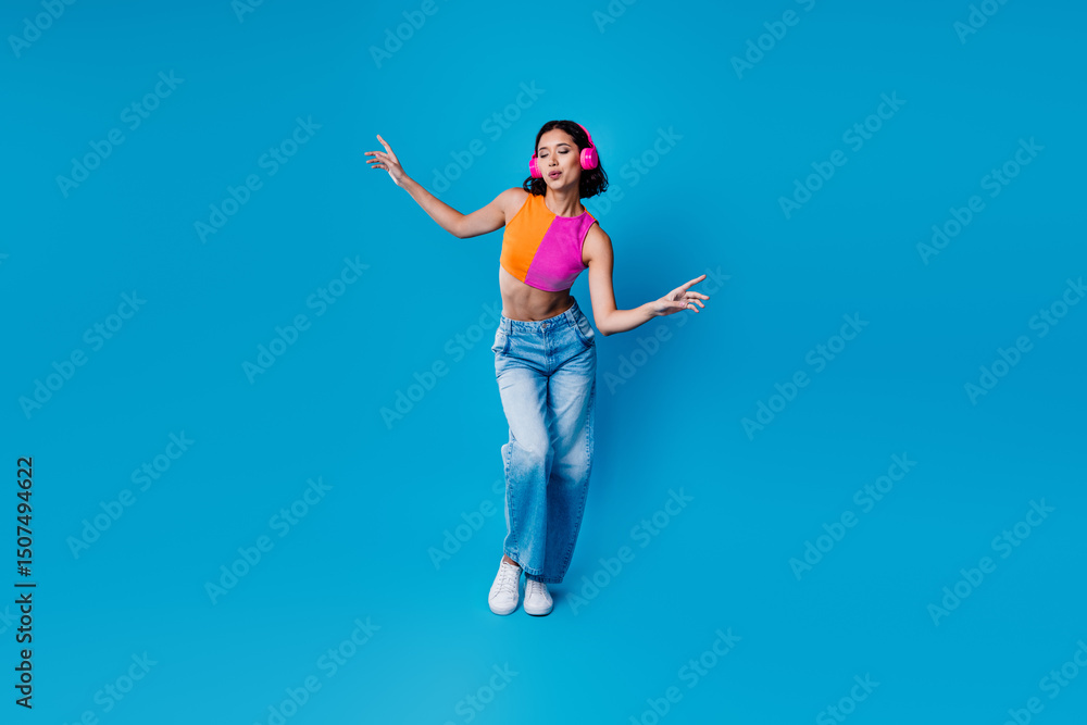 Young stylish woman dancing to music on blue backdrop wearing casual jeans and chic top with trendy headphones Young stylish woman dancing to music on blue backdrop wearing casual jeans and chic top with trendy headphones