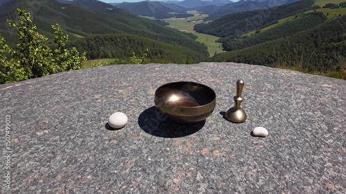 Singing bowl with striker and white stones on flat rock overlooking lush green valley
