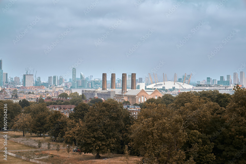 Fototapeta premium Panoramic view of London's skyline featuring the O2 Arena and Greenwich Power Station, with a lush park and modern cityscape under a cloudy sky.