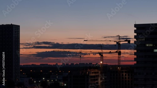 Time lapse. Urban landscape at night time. Construction site with silhouettes of two working cranes on the sunset urban skyline background