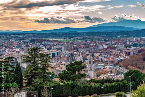 Panoramic view of Girona at sunset with church towers, historic and modern architecture, and the Pyrenees mountains in the background. The cityscape glows under a dramatic evening sky, Spain