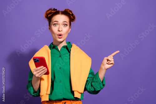 Fotografía Cheerful red haired woman holding a smartphone while gesturing amazedly, standin