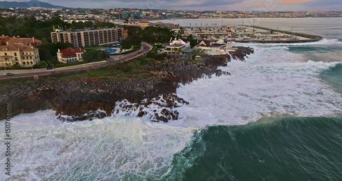 Aerial view hugging rocky coast by the sea in Cascais, Portugal with waves crashing underneath