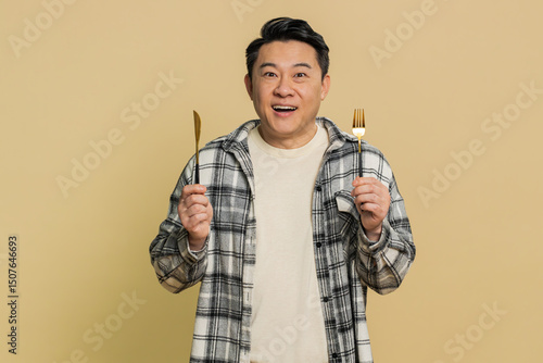 Ready to eat. Hungry Asian Chinese middle-aged man waiting for serving dinner dishes with with restlessness holding cutlery fork knife, will appreciate delicious restaurant meal on beige background