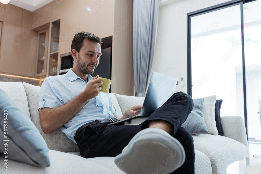 A man enjoys a relaxing moment at home, sipping coffee and working on his laptop while lounging on a comfortable sofa in a modern, bright living space with natural light and calm surroundings. A man enjoys a relaxing moment at home, sipping coffee and working on his laptop while lounging on a comfortable sofa in a modern, bright living space with natural light and calm surroundings.