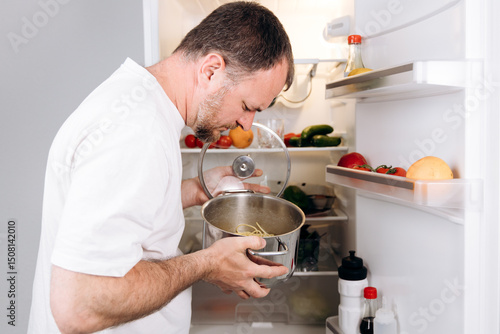 Man inspecting a pot of spoiled food in an open refrigerator filled with various vegetables and fruits, highlighting the importance of food safety and proper storage practices in a kitchen environment