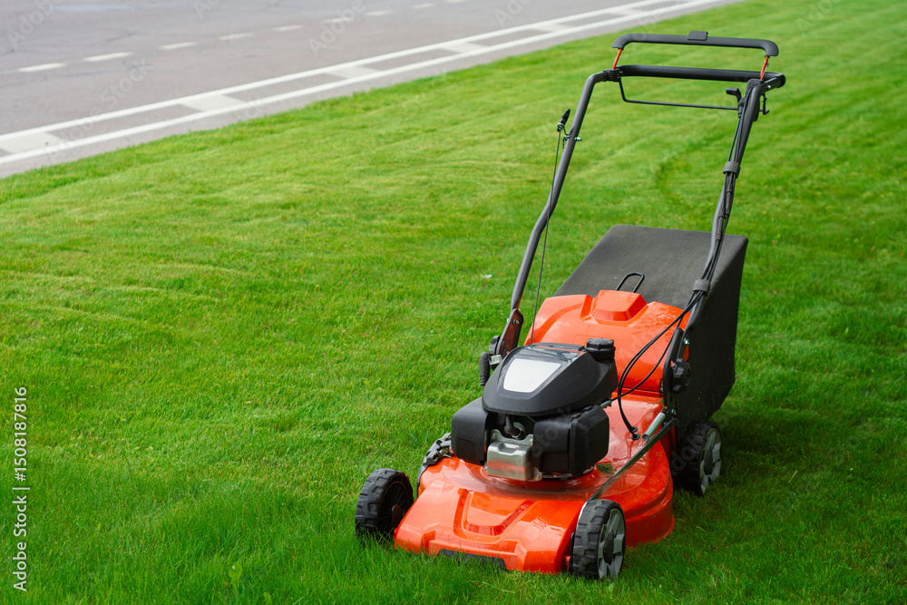 Fototapeta premium Bright orange lawn mower on lush green grass. Concept garden care, landscaping and summer outdoor work. Garden tool for lawn care and country lifestyle.