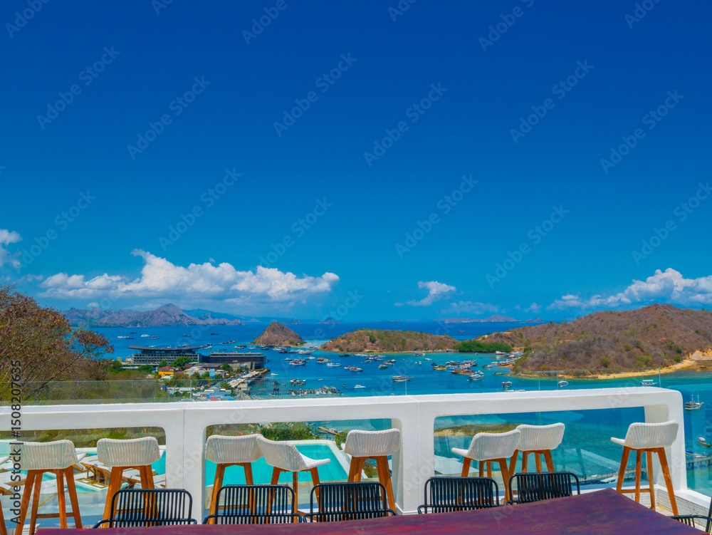 Fototapeta premium An outdoor dining area with a wooden table and chairs, overlooking the sea with mountains in the distance, under a clear blue sky. This serene setting is perfect for enjoying Labuan Bajo’s beauty