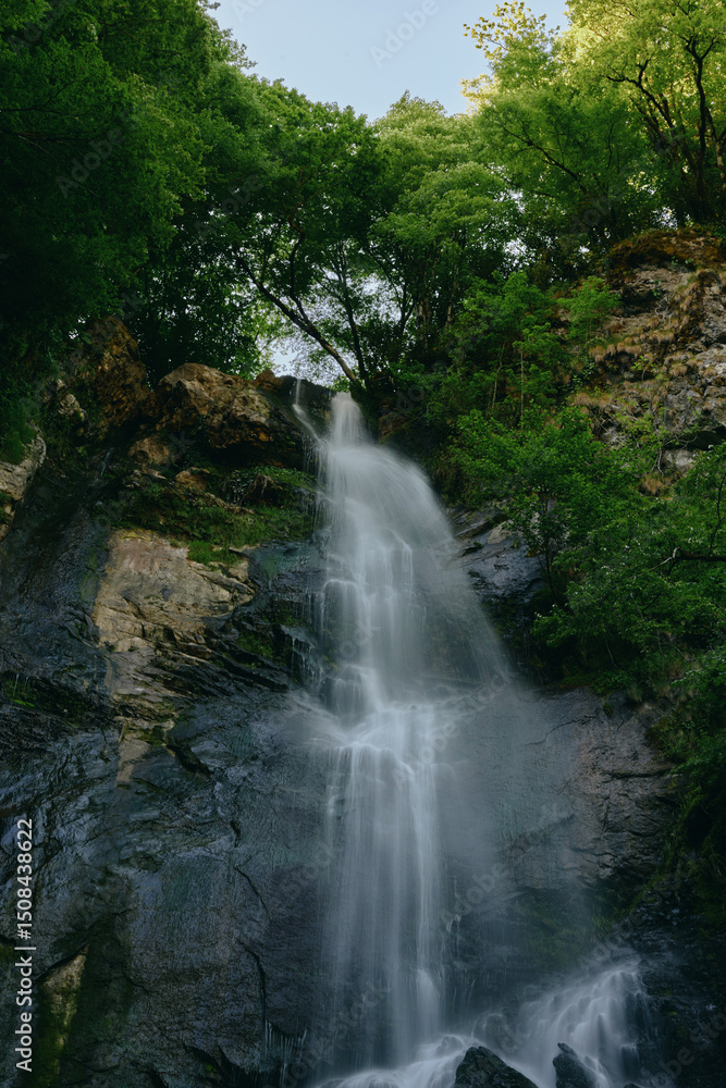 Obraz premium Natural waterfall cascading down rocky cliff surrounded by lush green forest, clear flowing water, peaceful outdoor landscape with vibrant foliage and tranquil atmosphere.