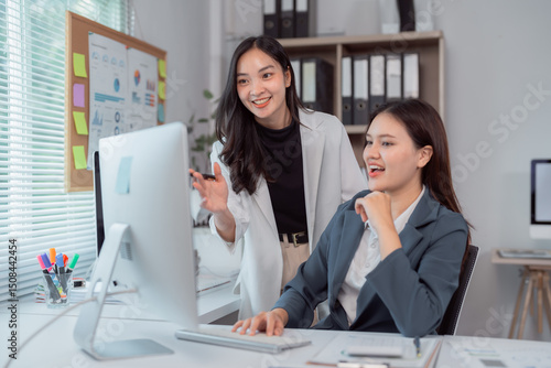 Two asian businesswomen are discussing a project while looking at desktop computer in a modern office, collaborating on a task and sharing ideas