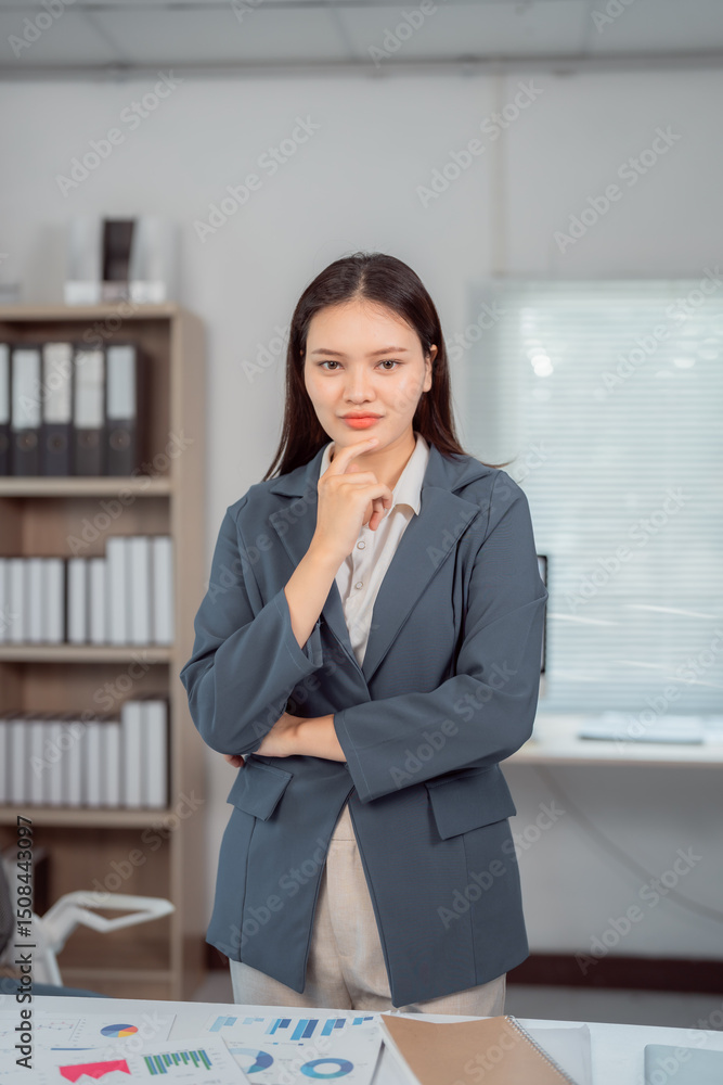 Young asian businesswoman in suit is thinking about marketing strategy while standing in office with documents on table and bookshelf in background Young asian businesswoman in suit is thinking about marketing strategy while standing in office with documents on table and bookshelf in background