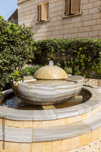A modern marble fountain stands in front of a stone building with green bushes.