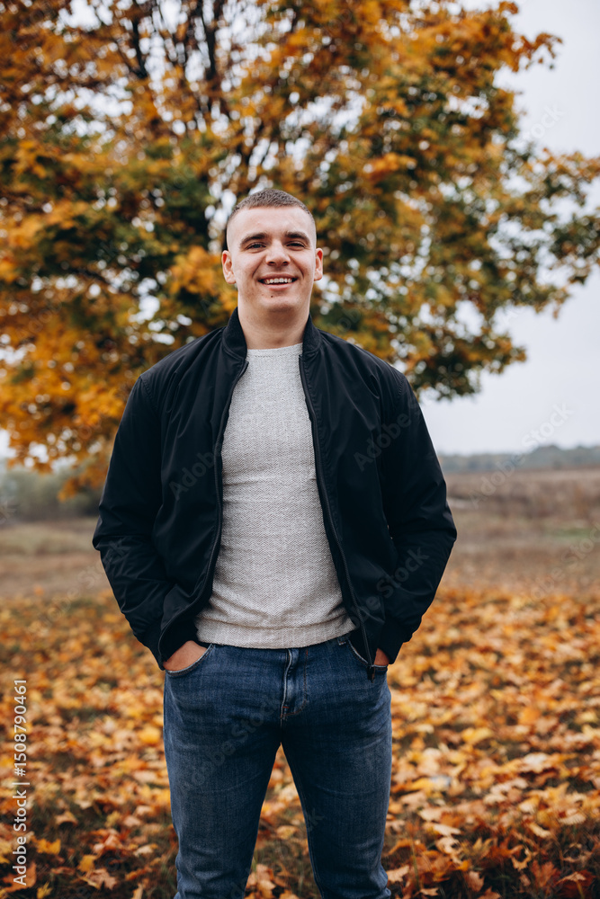 Smiling young man standing outdoors in an autumn park. He is wearing a black jacket and jeans, posing confidently in front of a tree with vibrant orange and yellow leaves. Casual and cheerful mood. Smiling young man standing outdoors in an autumn park. He is wearing a black jacket and jeans, posing confidently in front of a tree with vibrant orange and yellow leaves. Casual and cheerful mood.