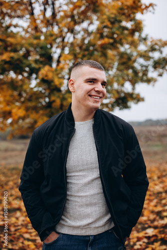Smiling young man standing outdoors in an autumn park. He is wearing a black jacket and jeans, posing confidently in front of a tree with vibrant orange and yellow leaves. Casual and cheerful mood.