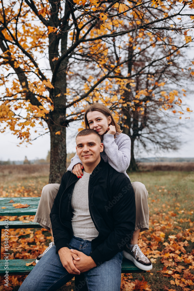 Young couple sitting and hugging on a bench under a yellow-leaved tree. Autumn idyll in nature, a romantic moment among fallen leaves in a field. A warm, cozy atmosphere surrounded by colorful leaves Young couple sitting and hugging on a bench under a yellow-leaved tree. Autumn idyll in nature, a romantic moment among fallen leaves in a field. A warm, cozy atmosphere surrounded by colorful leaves