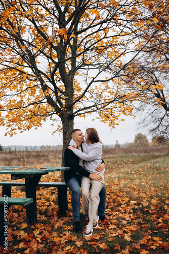 Young couple sitting and hugging on a bench under a yellow-leaved tree. Autumn idyll in nature, a romantic moment among fallen leaves in a field. A warm, cozy atmosphere surrounded by colorful leaves 