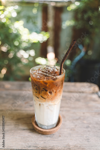 Glass of iced latte with milk and espresso layers served on a wooden table in a garden setting with lush green background and soft sunlight.