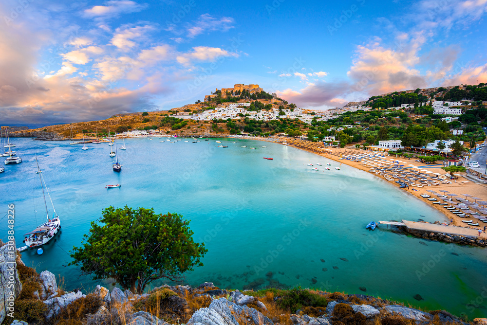 Naklejka premium Lindos city, Rhodes island, Greece: Panoramic view of Lindos bay at sunset with whitewashed village and the Acropolis, at Aegean Sea